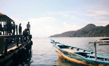 A group of people stands on a pier overlooking a moored boat by a scenic coastline under a clear sky.