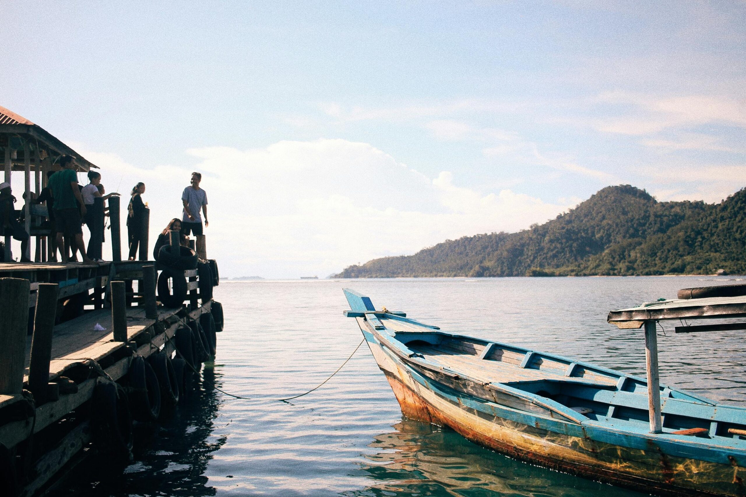 A group of people stands on a pier overlooking a moored boat by a scenic coastline under a clear sky.