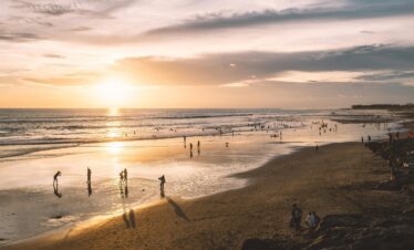 Silhouettes of faceless tourists on sandy coast near wavy ocean under cloudy sky at sunset