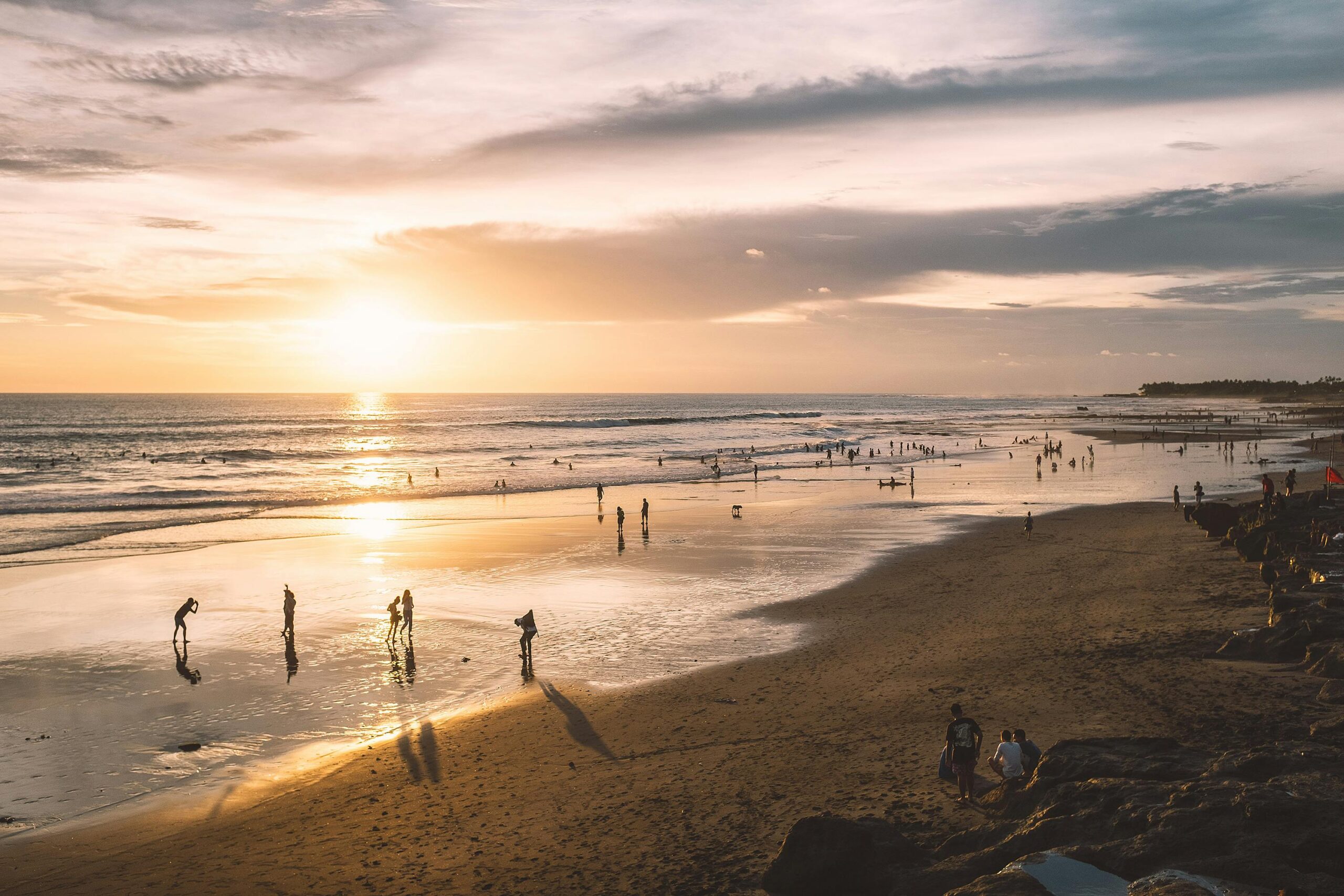 Silhouettes of faceless tourists on sandy coast near wavy ocean under cloudy sky at sunset